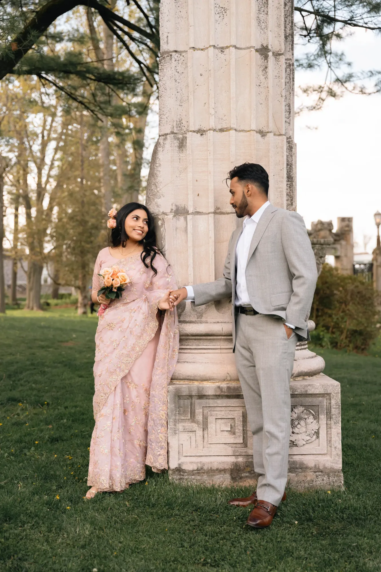 Bride-to-be in a blush pink embroidered saree holding flowers, hand in hand with her partner in a grey suit beside a stone column in a garden