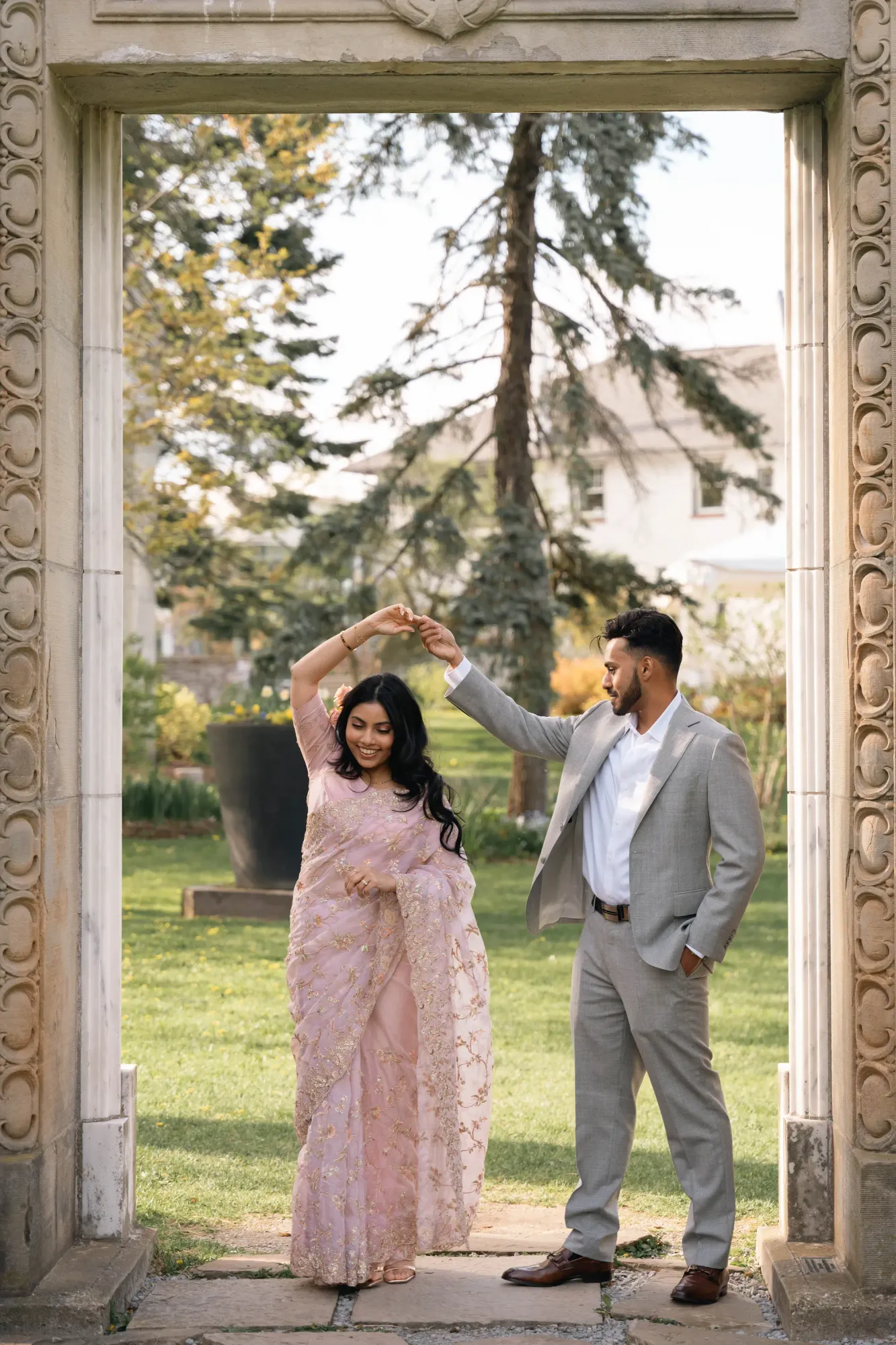 Couple dancing inside a carved stone doorway in a garden, the woman in a pink saree and the man in a grey suit