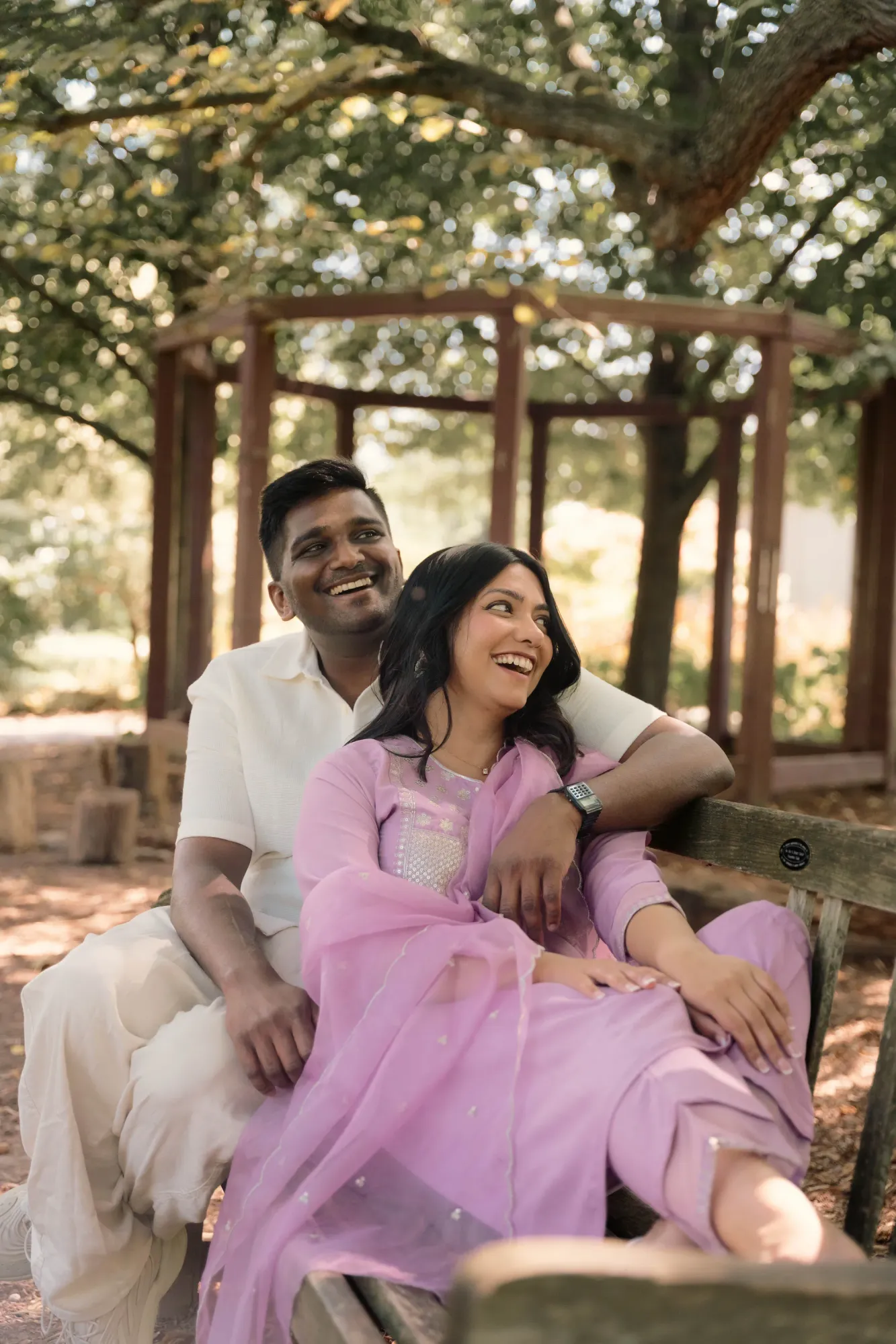 Laughing couple on a park bench, the woman in a lilac shalwar kameez with dupatta and the man in a cream outfit