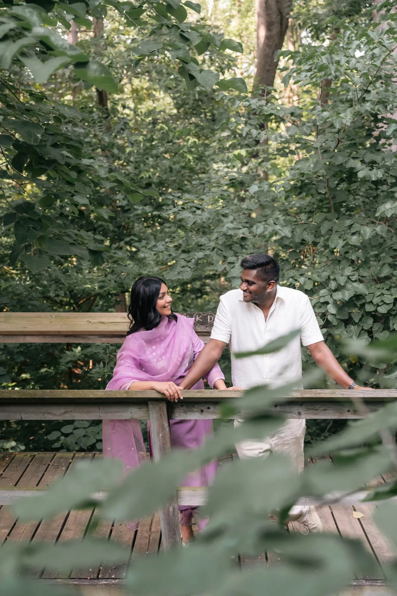 Couple leaning on a wooden railing in a leafy garden, the woman in a lilac shalwar kameez and the man in white