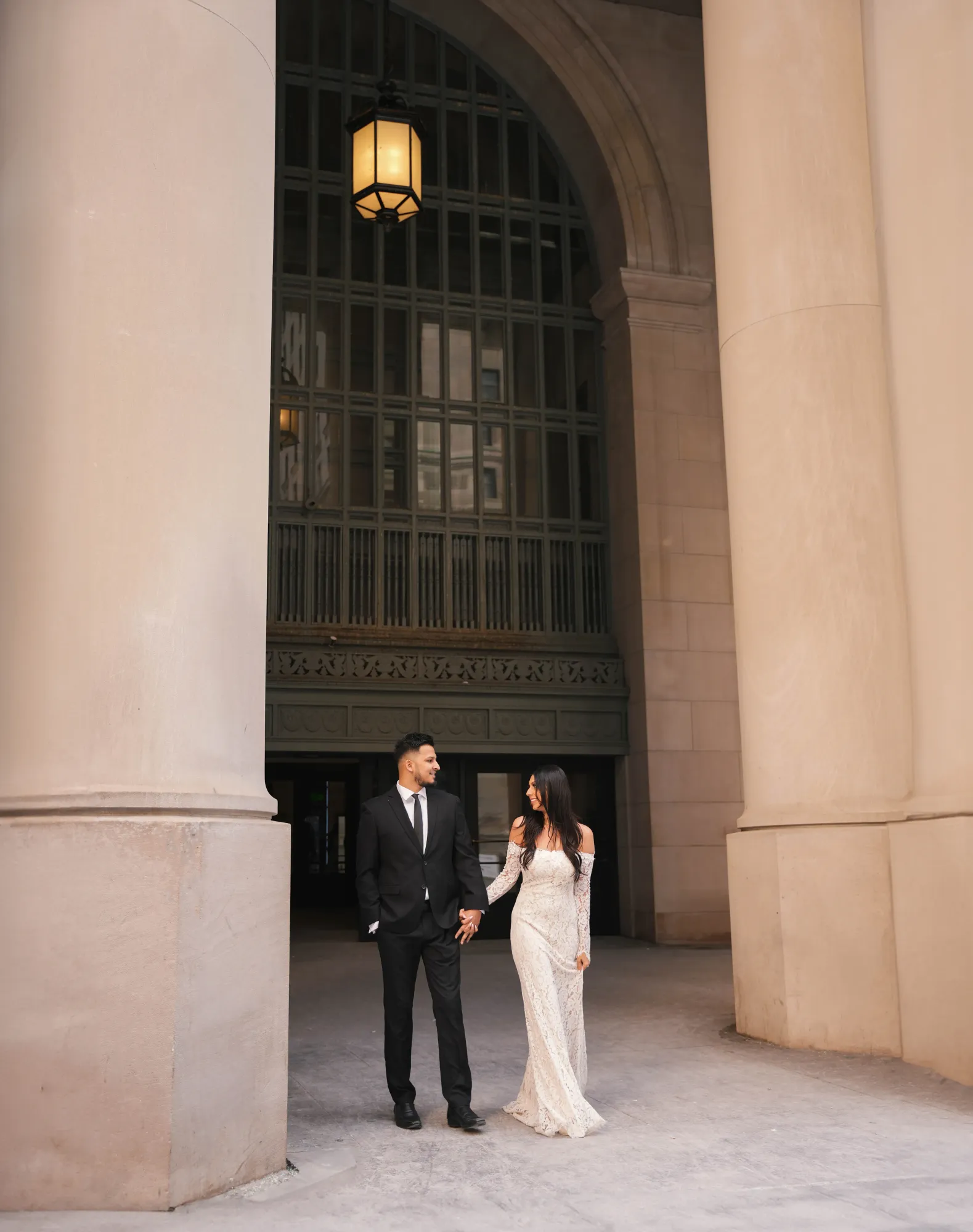 Bride in a long-sleeve lace gown and groom in a black suit holding hands under the stone arches of Union Station, Toronto