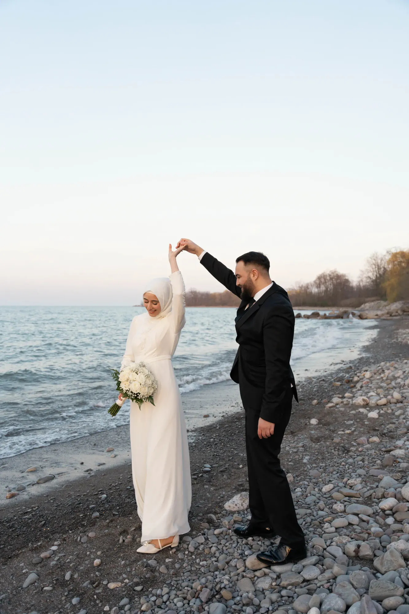 Groom twirling a bride in a white hijab and white gown on a pebble beach by the lake at sunset